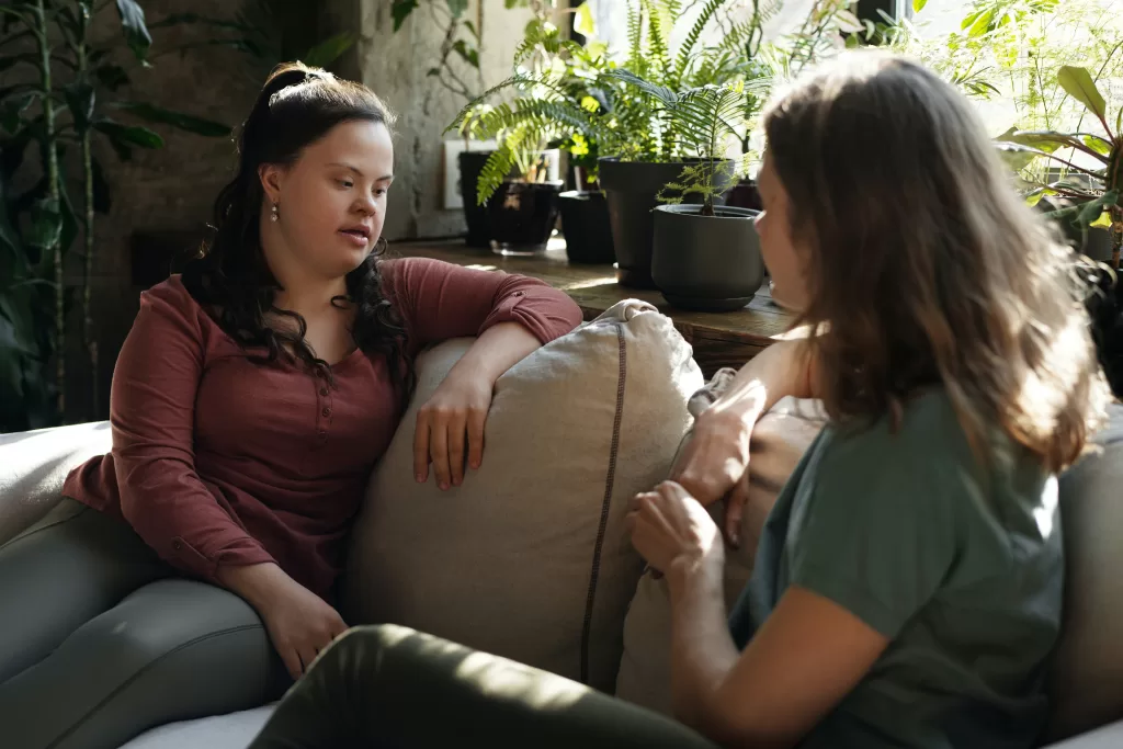 Two women sit on a couch having a conversation together. 