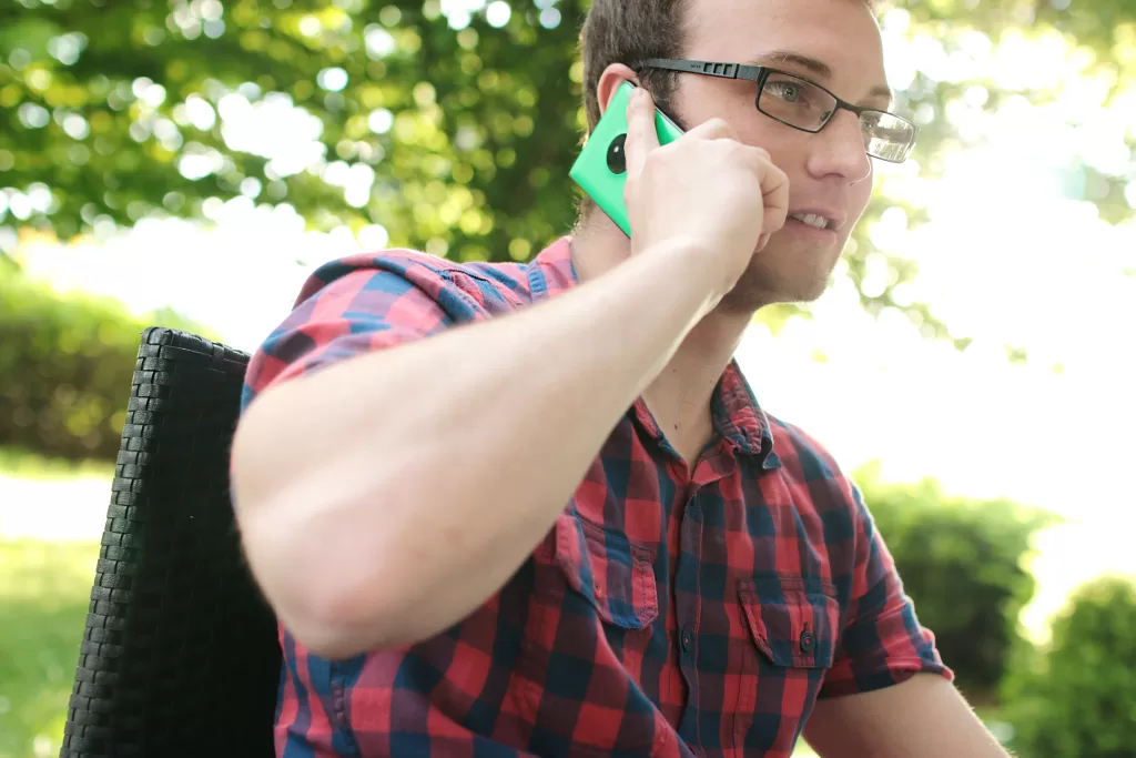 A man on the phone outside near a tree. He is wearing a red checkered shirt