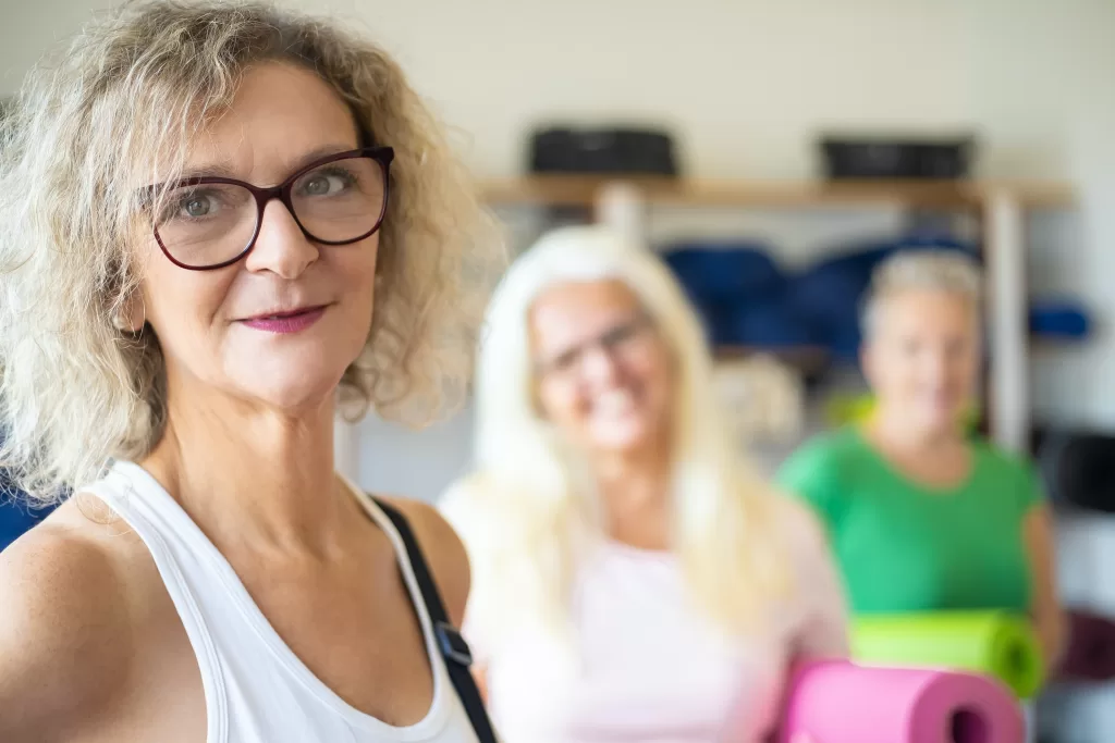 A white woman with blonde hair and glasses has a half smile. There are other women behind her with yoga mats in hand
