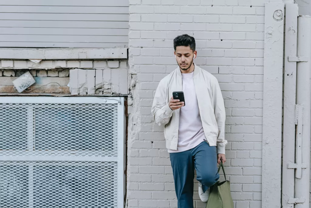 A young man is leaning a against a wall while looking at his phone.