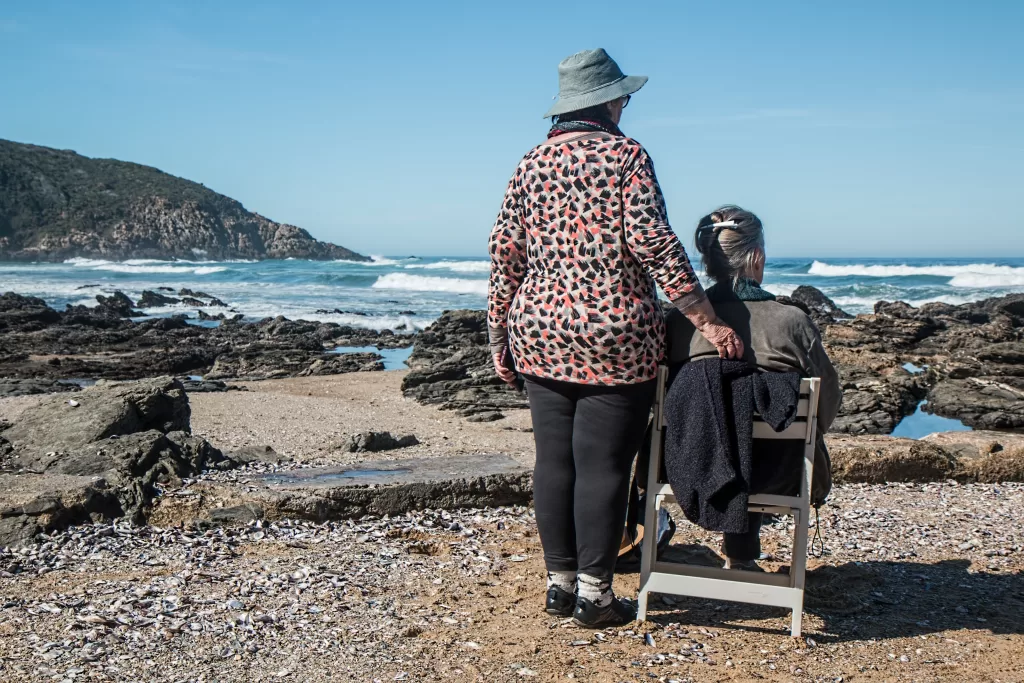 Two older women are on the beach. One is sitting on a chair and they are looking out at the ocean.