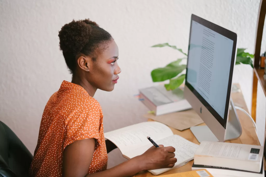 A young brown woman is writing on a notepad and looking at the computer