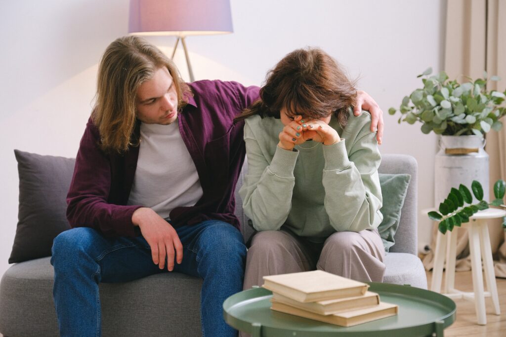 Brother supporting sister sits on couch with his arm around her while she rests her head in her hands