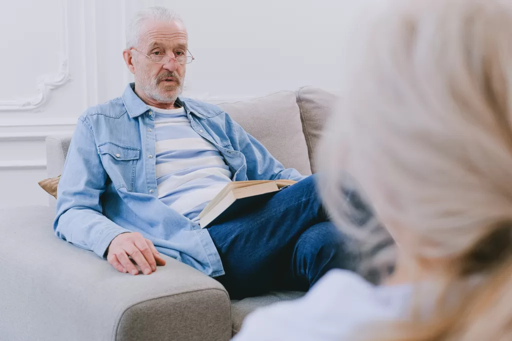 An older man sits on a couch opposite his wife. He has a book open on his lap.